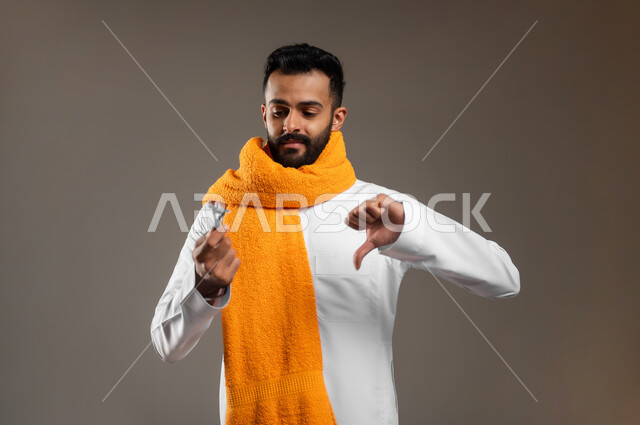 الحفاظ على الأناقة والنظافة الشخصية، الإشارة بعلامة عدم القبول والجودة السيئة، العناية بالبشرة والاهتمام بالمظهر الخارجي، بورتريه مقرب لرجل عربي خليجي سعودي يرتدي الثوب التقليدي يضع على عنقه المنشفة البرتقالية ويمسك بيده شفرة الحلاقة الرجالية، خلفية رمادية