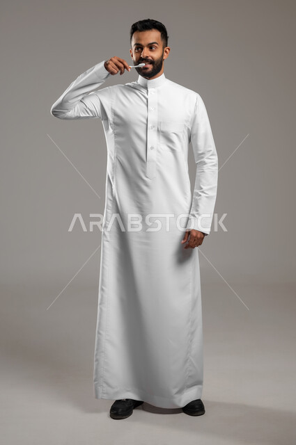 تجنب التسوس وأمراض اللثة، مفهوم العناية والإهتمام بصحة الأسنان، روتين نظافة الفم اليومية، بورتريه لرجل عربي خليجي سعودي يرتدي الثوب التقليدي يقوم بتنظيف أسنانه باستخدام الفرشاة، الجسم بالطول الكامل، خلفية رمادية