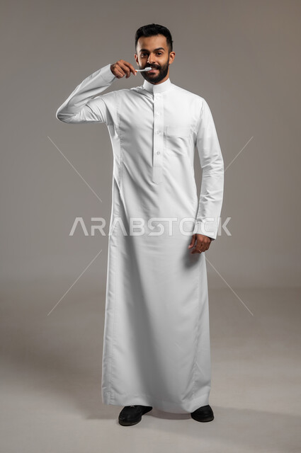 روتين نظافة الفم اليومية، النظر الى الكاميرا بإيماءات السعادة والسرور، بورتريه لرجل عربي خليجي سعودي يرتدي الثوب التقليدي يقوم بتنظيف أسنانه باستخدام الفرشاة، مفهوم العناية والإهتمام بصحة الأسنان، تجنب التسوس وأمراض اللثة، الجسم بالطول الكامل، خلفية رمادية