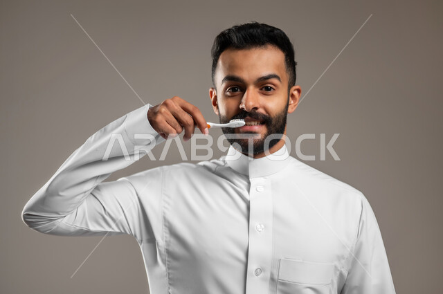 النظر الى الكاميرا بإيماءات السعادة والسرور، روتين نظافة الفم اليومية، بورتريه مقرب لرجل عربي خليجي سعودي يرتدي الثوب التقليدي يقوم بتنظيف أسنانه باستخدام الفرشاة، مفهوم العناية والإهتمام بصحة الأسنان، تجنب التسوس وأمراض اللثة، خلفية رمادية