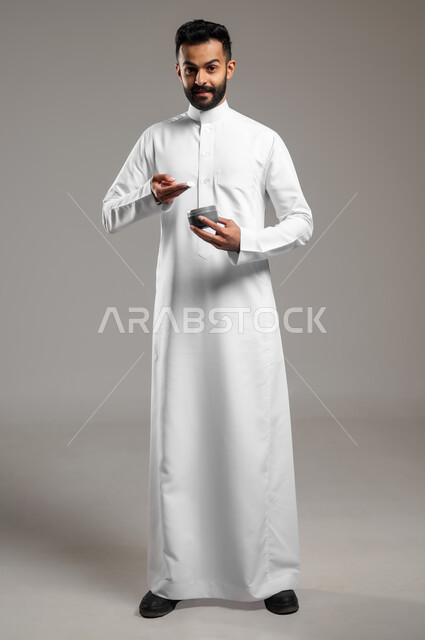 النظر للكاميرا بإيماءات السرور والسعادة، استخدام كريمات ومكونات تجميلية شبابية، مفهوم العناية والإهتمام بنضارة وتحسين البشرة، ترطيب وتنظيف وتفتيح الجلد، بورتريه لشاب عربي خليجي سعودي يرتدي الثوب التقليدي يمسك بيده كريم المرطب، الجسم بالطول الكامل، خلفية رمادية