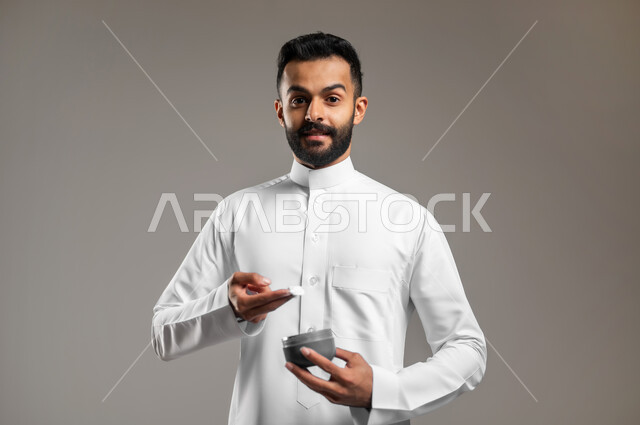 مفهوم العناية والإهتمام بنضارة وتحسين البشرة، النظر الى الكاميرا بإيماءات السعادة والسرور، استخدام كريمات ومكونات تجميلية شبابية، ترطيب وتنظيف وتفتيح الجلد، بورتريه مقرب لرجل عربي خليجي سعودي يرتدي الثوب التقليدي يمسك بيده كريم المرطب، خلفية رمادية