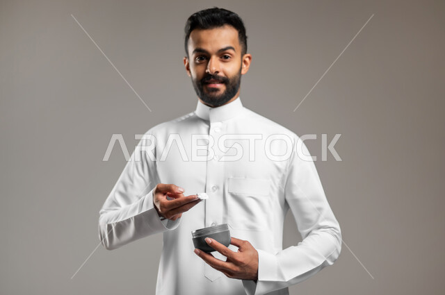 النظر الى الكاميرا بإيماءات السعادة والسرور، استخدام كريمات ومكونات تجميلية شبابية، مفهوم العناية والإهتمام بنضارة وتحسين البشرة، ترطيب وتنظيف وتفتيح الجلد، بورتريه مقرب لشاب عربي خليجي سعودي يرتدي الثوب التقليدي يمسك بيده كريم المرطب، خلفية رمادية