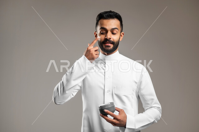 مفهوم العناية والإهتمام بنضارة وتحسين البشرة، ترطيب وتنظيف وتفتيح الجلد، بورتريه مقرب لشاب عربي خليجي سعودي يرتدي الثوب التقليدي يمسك بيده كريم المرطب ويضعه على الوجه، استخدام مكونات تجميلية شبابية، خلفية رمادية
