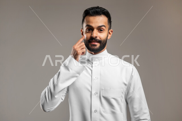 النظر الى الكاميرا بإيماءات السعادة والسرور، ترطيب وتنظيف وتفتيح الجلد، بورتريه مقرب لشاب عربي خليجي سعودي يرتدي الثوب التقليدي يضع كريم المرطب على الوجه، مفهوم العناية والإهتمام بنضارة وتحسين البشرة، استخدام مكونات تجميلية شبابية، خلفية رمادية