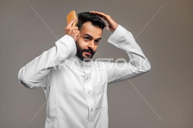 ترتيب وتمشيط الشعر، مفهوم العناية والاهتمام بالجمال والأناقة والمظهر الخارجي، بورتريه مقرب لشاب وسيم عربي خليجي سعودي يرتدي الثوب التقليدي يقوم بتصفيف شعره باستخدام فرشاة ( المشط )، خلفية رمادية