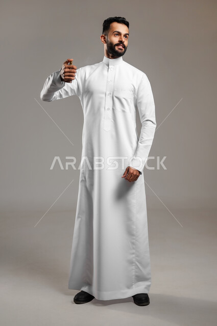 عطورات خليجية سعودية, استخدام الزيوت العطرية الطبيعية, مفهوم الاناقة والاهتمام بالمظهر الخارجي, بورتريه لشاب وسيم عربي خليجي سعودي يرتدي الثوب التقليدي يقوم بوضع العطر, التسويق لمنتجات العطور, الجسم بالطول الكامل، خلفية رمادية