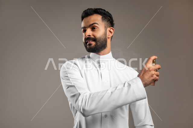 استخدام الزيوت العطرية الطبيعية, عطورات خليجية سعودية, مفهوم الاناقة والاهتمام بالمظهر الخارجي, بورتريه مقرب لشاب وسيم عربي خليجي سعودي يرتدي الثوب التقليدي يقوم بوضع العطر, التسويق لمنتجات العطور, خلفية رمادية