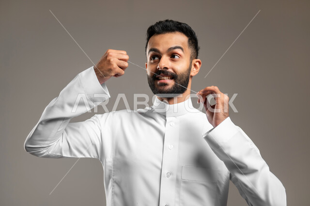 الاهتمام بالأناقة والمظهر الخارجي، المحافظة على ابتسامة بيضاء، بورتريه مقرب لشاب وسيم عربي خليجي سعودي يرتدي الثوب التقليدي يقوم بتنظيف أسنانه باستخدام الخيط السني، مفهوم العناية بصحة الفم والأسنان، خلفية رمادية