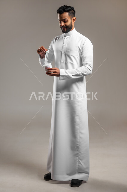 استخدام أعواد ونكاشات القطن، العناية والاهتمام بصحة الأذن، بورتريه لشاب وسيم عربي خليجي سعودي يرتدي الثوب التقليدي يمسك بيده نكاشة قطنية، روتين نظافة الأذنين اليومي، الجسم بالطول الكامل، خلفية رمادية