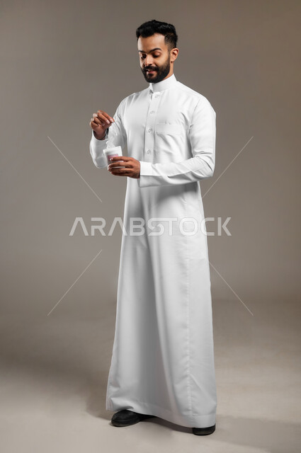 استخدام أعواد ونكاشات القطن، روتين نظافة الأذنين اليومي، العناية والاهتمام بصحة الأذن، بورتريه لشاب وسيم عربي خليجي سعودي يرتدي الثوب التقليدي يمسك بيده نكاشة قطنية، الجسم بالطول الكامل، خلفية رمادية