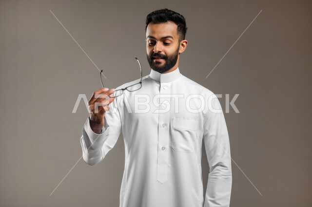 تحسين الرؤية ومحاولة تعديل البصر، الاناقة والاهتمام بالمظهر الخارجي، بورتريه مقرب لشاب وسيم عربي خليجي سعودي يرتدي الثوب التقليدي يمسك بيده نظارة طبية وينظر إليها، ارتداء النظارات الخاصة بضعف النظر، خلفية رمادية