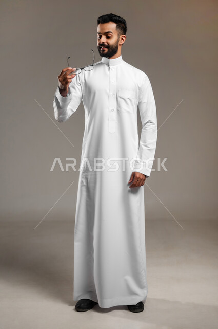 الاناقة والاهتمام بالمظهر الخارجي، بورتريه لشاب وسيم عربي خليجي سعودي يرتدي الثوب التقليدي يمسك بيده نظارة طبية وينظر إليها، ارتداء النظارات الخاصة بضعف النظر، تحسين الرؤية ومحاولة تعديل البصر، الجسم بالطول الكامل، خلفية رمادية