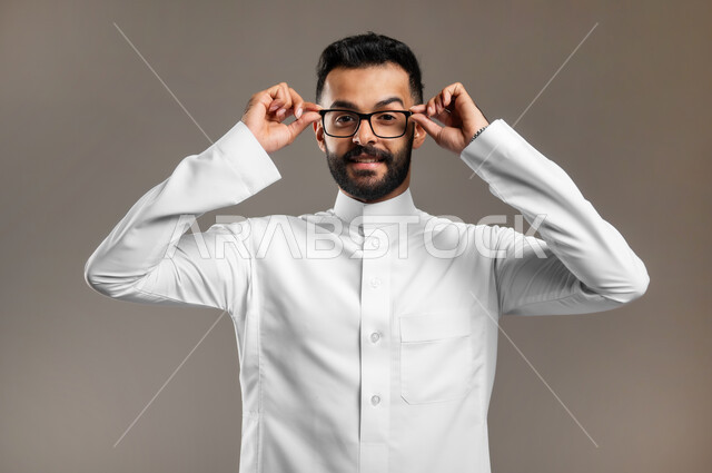 Improving vision and trying to adjust eyesight, taking care of external appearance, wearing glasses for low vision, close-up portrait of a handsome Saudi Gulf Arab young man wearing a traditional dress and wearing glasses, looking at the camera with happy gestures, gray background