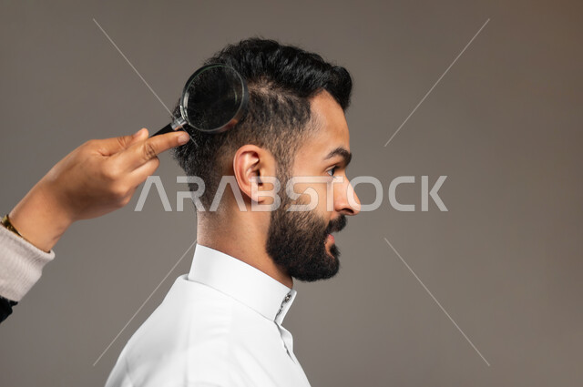 فحص فروة الرأس، سلامة الشعر من القشرة والقمل، المحافظة على النظافة والمظهر الخارجي، بورتريه مقرب ليد تضع العدسة المكبرة على رأس شاب وسيم عربي سعودي خليجي يرتدي الثوب التقليدي، خلفية رمادية