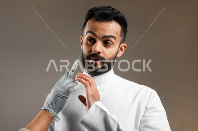 الاهتمام بالمظهر الخارجي، إخفاء تجاعيد الوجه، مكافحة الشيخوخة المبكرة، مفهوم العناية بنضارة وتحسين البشرة، بورتريه مقرب ليد أخصائي جلدية يقوم بحقن مادة البوتوكس على وجه شاب وسيم عربي خليجي سعودي يرتدي الثوب التقليدي، رفع اليد بتعابير الحذر والخوف، خلفية رمادية