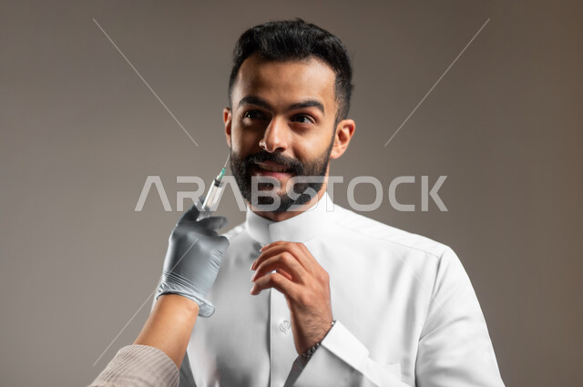 مكافحة الشيخوخة المبكرة، العناية والإهتمام بنضارة وتحسين البشرة، بورتريه مقرب ليد أخصائي جلدية يقوم بحقن مادة البوتوكس على وجه شاب وسيم عربي خليجي سعودي يرتدي الثوب التقليدي، إخفاء تجاعيد الوجه، رفع اليد بتعابير الحذر والخوف، خلفية رمادية