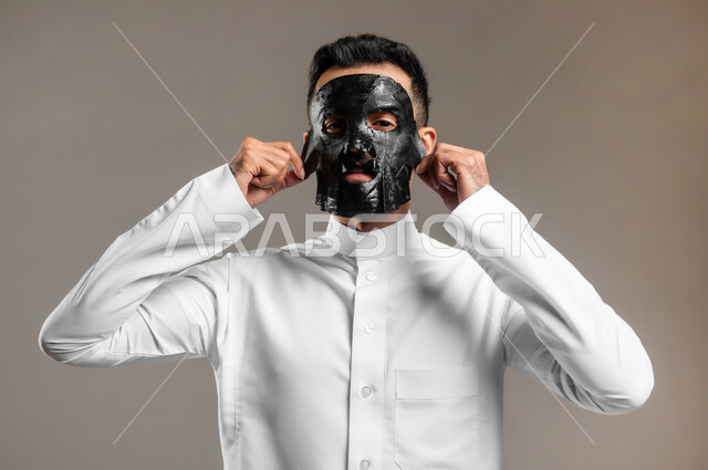 مفهوم العناية والاهتمام بالمظهر الخارجي، استخدام مكونات تجميلية، ماسك تنظيف وتقشير البشرة، بورتريه مقرب لشاب وسيم عربي خليجي سعودي يرتدي الثوب التقليدي يقوم بنزع  قناع الفحم الاسود من على وجهه للتخلص من الرؤوس السوداء، خلفية رمادية