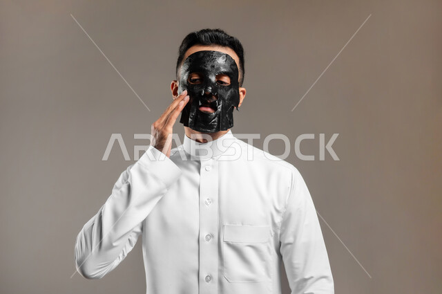 ماسك تنظيف وتقشير البشرة، استخدام مكونات تجميلية، بورتريه مقرب لشاب وسيم عربي خليجي سعودي يرتدي الثوب التقليدي يقوم بوضع قناع الفحم الاسود على وجهه للتخلص من الرؤوس السوداء، مفهوم العناية والاهتمام بالمظهر الخارجي، خلفية رمادية