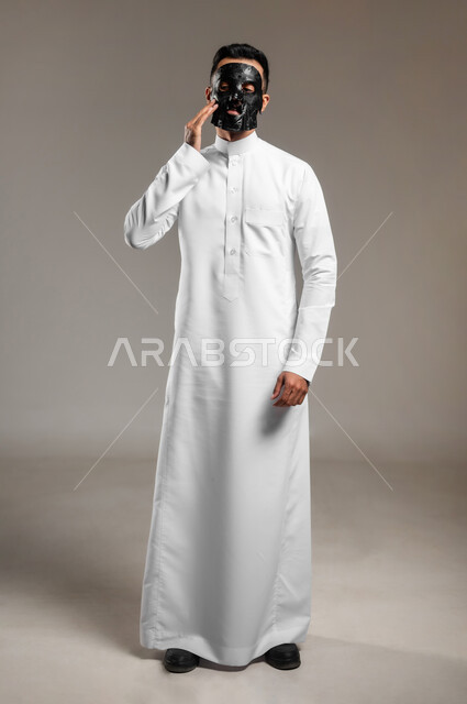 استخدام مكونات تجميلية، ماسك تنظيف وتقشير البشرة، بورتريه لشاب وسيم عربي خليجي سعودي يرتدي الثوب التقليدي يقوم بوضع قناع الفحم الاسود على وجهه للتخلص من الرؤوس السوداء، مفهوم العناية والاهتمام بالمظهر الخارجي، الجسم بالطول الكامل، خلفية رمادية