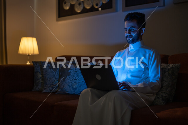 استعمال الأجهزة والتقنيات الحديثة والمتطورة, البحث عن شيء ما عبر الانترنت، تصفح مواقع التواصل الاجتماعي، متابعة الأفلام والمسلسلات المفضلة، إنجاز مهام العمل عن بعد, رجل عربي خليجي سعودي يجلس على أريكة في غرفة المعيشة في وقت الليل و يحمل جهاز الحاسوب المحمول