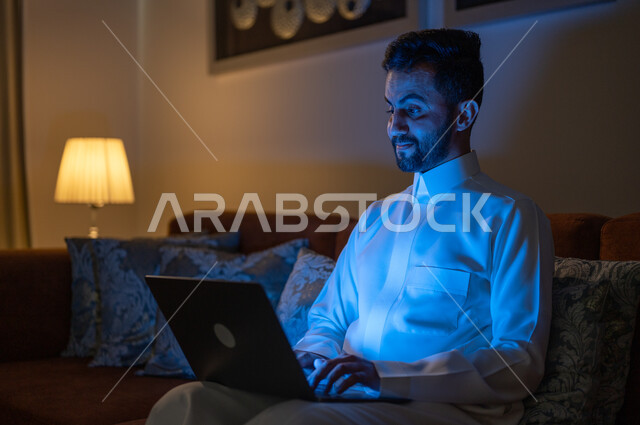 Using modern and advanced devices and technologies, searching for something online, browsing social networking sites, following favorite movies and series, completing work tasks remotely, a Saudi Gulf Arab man sitting on a sofa in the living room at night time and holding a laptop