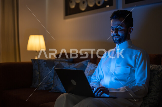 البحث عن شيء ما عبر الانترنت، تصفح مواقع التواصل الاجتماعي، متابعة الأفلام والمسلسلات المفضلة، إنجاز مهام العمل عن بعد, تطور الأجهزة والتقنيات الحديثة, رجل عربي خليجي سعودي يجلس على أريكة في غرفة المعيشة في وقت الليل و يحمل جهاز الحاسوب المحمول