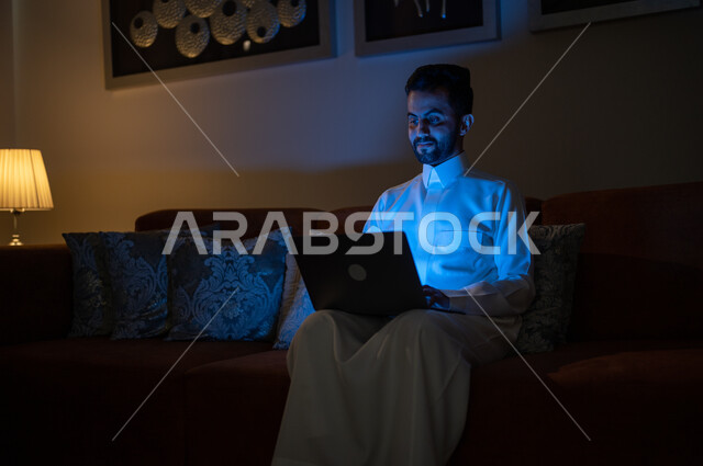 متابعة الأفلام والمسلسلات المفضلة، إنجاز مهام العمل عن بعد, تطور الأجهزة والتقنيات الحديثة, تصفح مواقع التواصل الاجتماعي، البحث عن شيء ما عبر الانترنت، رجل عربي خليجي سعودي يجلس على أريكة في غرفة المعيشة في وقت الليل و يحمل جهاز الحاسوب المحمول
