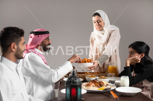 ايماءات السعادة  بقدوم الشهر الفضيل, توزيعات و ضيافة تمورالمملكة العربية السعودية, بورتريه مقرب لامرأة عربية خليجية سعودية تقوم بتقديم طبق من التمر السعودي لعائلتها على مائدة الطعام, الاحتفال بقدوم الشهر القضيل, خلفية رمادية