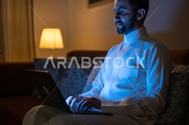 Completing work tasks remotely, the development of modern devices and technologies, following social networking sites, watching favorite movies and series, searching for something online, a Saudi Gulf Arab man sitting on a sofa in the living room at night and holding a laptop.