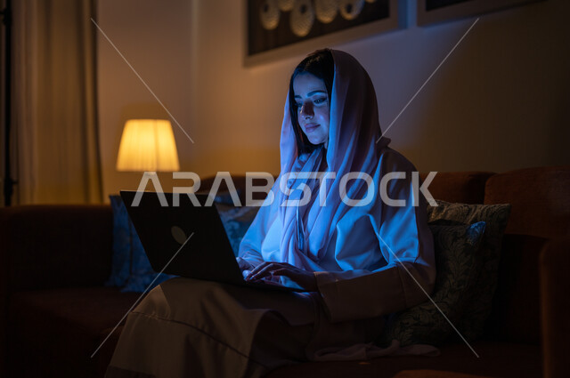 تطور الأجهزة والتقنيات الحديثة, متابعة مواقع التواصل الاجتماعي، إنجاز مهام العمل عن بعد, مشاهدة الأفلام والمسلسلات المفضلة، البحث عن شيء ما عبر الانترنت، امرأة عربية خليجية سعودية تجلس على أريكة في غرفة المعيشة في وقت الليل و تحمل جهاز الحاسوب المحمول