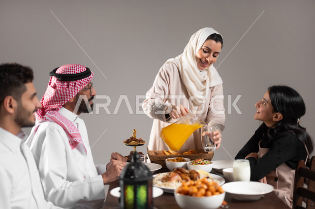توزيع العصير الى افراد العائلة على مائدة الطعام, بورتريه  لعائلة عربية خليجية سعودية مجتمعة على طاولة الطعام, العصائر و الاصناف الشرقية اللذيذة, تنظيم سفرة فطور رمضان, ايماءات السعادة  بقدوم الشهر الفضيل, خلفية رمادية
