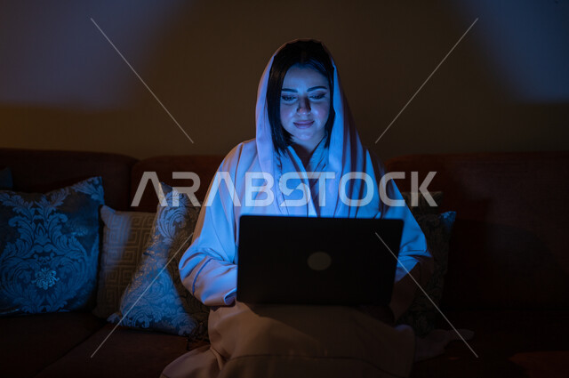 مراقبة مواقع التواصل الاجتماعي، تطور الأجهزة والتقنيات الحديثة, إنجاز مهام العمل عن بعد, مشاهدة الأفلام والمسلسلات المفضلة، البحث عن شيء ما عبر الانترنت، امرأة عربية خليجية سعودية تجلس على أريكة في غرفة المعيشة في وقت الليل و تحمل جهاز الحاسوب المحمول