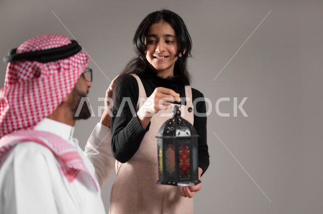 فرحة الابناء بقدوم الشهر المبارك, بورتريه مقرب لابنة عربية سعودية مبتسمة تحمل فانوس رمضان و تشارك والدها فرحتها بقدوم رمضان, الاحتفال مع الاهل بقدوم الشهر الفضيل, الاحتفال بالاعياد و المناسبات الاسلامية, خلفية رمادية