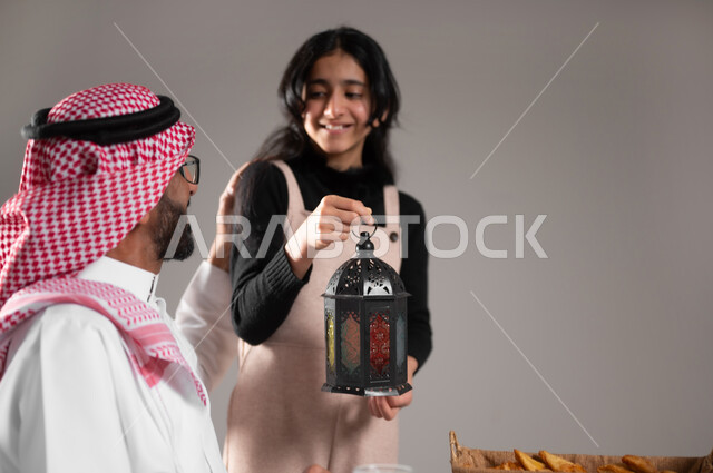 مشاركة الابناء فرحة قدوم الشهر المبارك, بورتريه مقرب لابنة عربية سعودية مبتسمة تحمل الفانوس الرمضاني و تقدمه لوالدها, تعظيم و تقديرالشهر الفضيل, الاحتفال بالاعياد و المناسبات الاسلامية, خلفية رمادية
