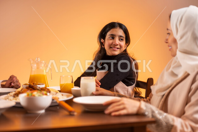 بناء علاقات صحيه سليمه مع الأولاد، قضاء أوقات ممتعه مع الأبناء، بورتريه مقرب لابنة عربيه خليجية سعوديه تجلس على طاوله الطعام المليئة بأشهى الاكلات و أم سعودية تقوم بتقديم العصير اللذيذ لابنتها، متعة لمة الاهل على السفرة, ايماءات السعادة بمناسبة قدوم الشهر الفضيل, خلفية برتقالية