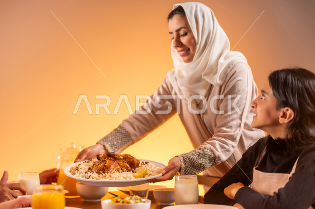 تجهيز طاولة الطعام المليئة بالمأكولات الشهية, بورتريه مقرب لأم عربية خليجية سعودية تقوم بتحضير سفرة رمضان و وضع الطبق الرئيسي عليها, المقبلات و الاصناف العربية الشرقية, ايماءات السعادة بمناسبة قدوم الشهر الفضيل, الاحتفال بالاعياد و المناسبات الاسلامية, خلفية برتقالية