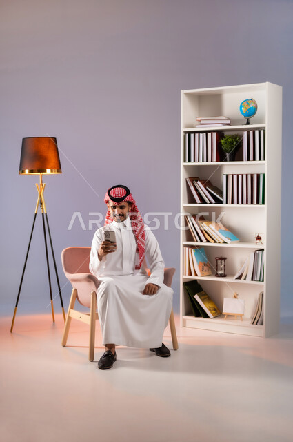 الاندماج بالتواصل مع الأصدقاء والأقارب عبر التطبيقات والتقنيات الحديثة والمتطورة, إجراء مكالمة فيديو بإيماءات السرور عبر الانترنت, بورتريه لرجل عربي خليجي سعودي مبتسم يرتدي الثوب التقليدي والشماغ يستخدم الهاتف المحمول, خلفية ملونة