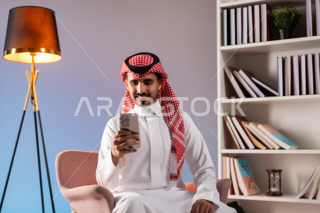 التواصل مع الأصدقاء والأقارب عبر التطبيقات والتقنيات الحديثة والمتطورة, إجراء مكالمة فيديو بإيماءات السرور عبر الانترنت, بورتريه لرجل عربي خليجي سعودي مبتسم يرتدي الثوب التقليدي والشماغ يستخدم الهاتف المحمول,  خلفية ملونة