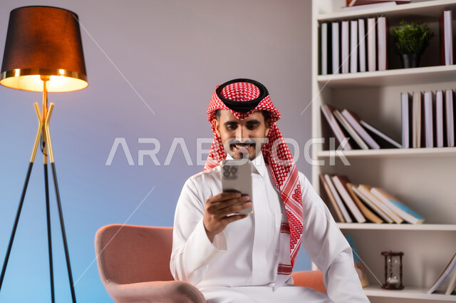 إجراء مكالمة فيديو بإيماءات السرور عبر الانترنت, بورتريه لرجل عربي خليجي سعودي مبتسم يرتدي الثوب التقليدي والشماغ يستخدم الهاتف المحمول, التواصل مع الأصدقاء والأقارب عبر التطبيقات والتقنيات الحديثة والمتطورة, خلفية ملونة
