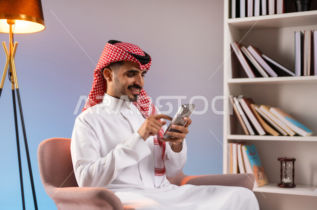 الدردشة والمحادثة عبر الونلاين, القيام بالبث المباشر عبر السوشال ميديا, بورتريه لرجل عربي خليجي سعودي مبتسم يرتدي الثوب التقليدي والشماغ يستخدم الهاتف المحمول بإيماءات السعادة, التواصل مع الأصدقاء والأقارب عبر التطبيقات والتقنيات الحديثة والمتطورة, خلفية ملونة