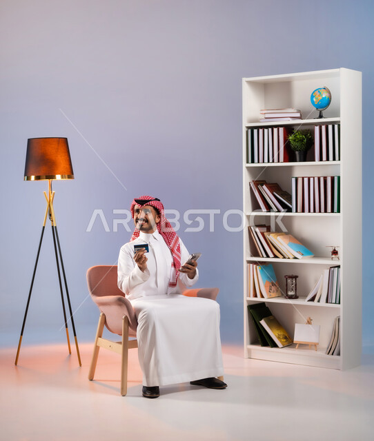 النظر إلى شي ما بإيماءات السرور, تصفح تطبيقات المصارف والبنوك عبر الجوال، الشراء والدفع عبر الاونلاين, استخدام بطاقة الإئتمان في التسوق الالكتروني, بورتريه لرجل عربي خليجي سعودي يرتدي الثوب التقليدي والشماغ يجلس على كرسي ويمسك البطاقة البنكية, خلفية ملونة