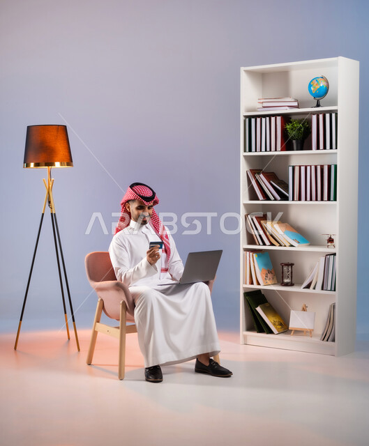 سهولة الشراء والدفع عن طريق بطاقة الإئتمان البنكية, إمكانية تسديد الفواتير بالبطاقات الإلكترونية, استخدام الأجهزة والتقنيات الحديثة، بورتريه لرجل عربي خليجي سعودي مبتسم يرتدي الثوب التقليدي والشماغ يمسك بيده بطاقة الصراف ويعمل على الحاسب المحمول, خلفية ملونة