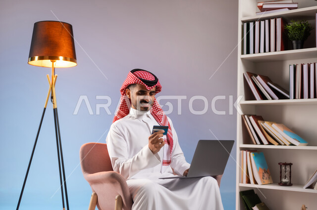 إمكانية تسديد الفواتير بالبطاقات الإلكترونية, استخدام الأجهزة والتقنيات الحديثة, سهولة الشراء والدفع عن طريق بطاقة الإئتمان البنكية، بورتريه لرجل عربي خليجي سعودي مبتسم يرتدي الثوب التقليدي والشماغ يمسك بيده بطاقة الصراف ويعمل على الحاسب المحمول, خلفية ملونة