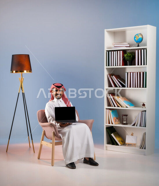 Using modern and advanced technical devices, managing business remotely, a portrait of a smiling Saudi Arabian Gulf man wearing a traditional thobe, displaying the laptop with a blank black screen, browsing electronic applications and programs through the laptop, colored background