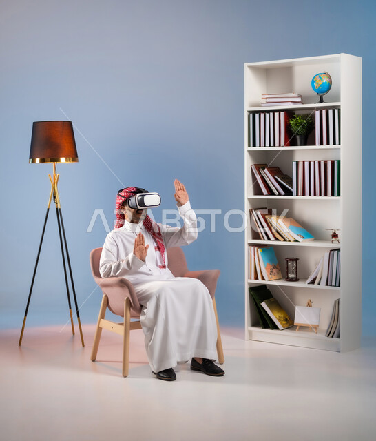 التفاعل والاندماج ومحاكاة العالم الخيالي, الترفيه والتسلية في وقت الفراغ, الاستمتاع بمشاهدة أفلام الواقع الافتراضي, بورتريه لرجل عربي خليجي سعودي يرتدي الثوب التقليدي ونظارة في آر ثلاثية الأبعاد يرفع يديه لمحاولة لمس شيء ما, خلفية ملونة
