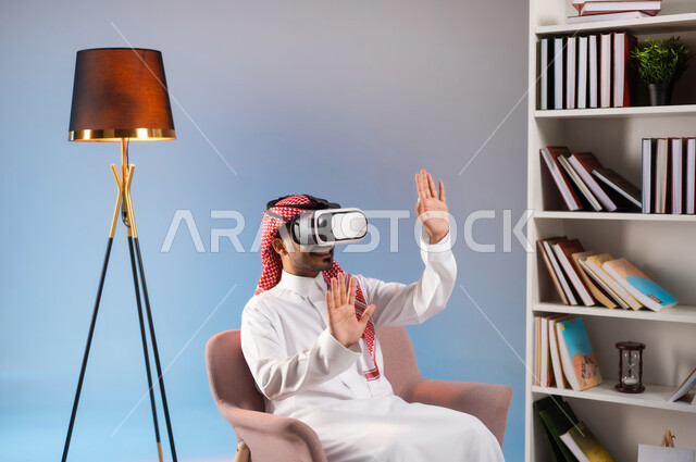 الترفيه والتسلية في وقت الفراغ, الاستمتاع بمشاهدة أفلام الواقع الافتراضي, بورتريه لرجل عربي خليجي سعودي يرتدي الثوب التقليدي ونظارة في آر ثلاثية الأبعاد يرفع يديه لمحاولة لمس شيء ما, التفاعل والاندماج ومحاكاة العالم الخيالي, خلفية ملونة
