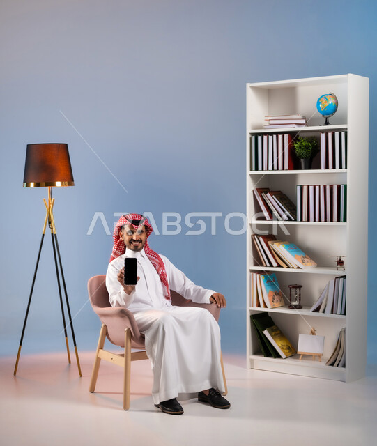 الجلوس والنظر إلى الكاميرا بإيماءات السرور, عرض تطبيقات وخدمات الهاتف المحمول, بورتريه لشاب عربي خليجي سعودي يرتدي الثوب التقليدي والشماغ يحمل الجوال بشاشة سوداء فارغة, استخدام الأجهزة والتقنيات الحديثة, التواصل مع الأهل والأصدقاء عبر الأونلاين, خلفية ملونة