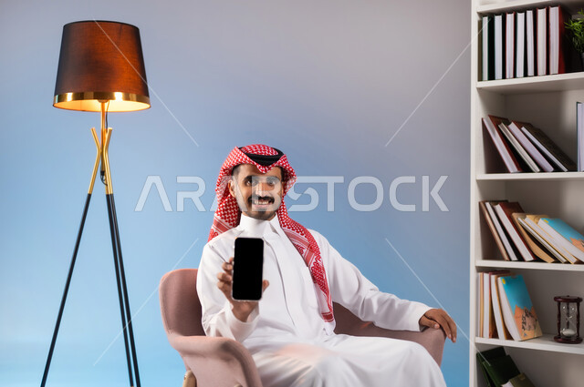 عرض تطبيقات وخدمات الهاتف المحمول, الجلوس والنظر إلى الكاميرا بإيماءات السرور, بورتريه لشاب عربي خليجي سعودي يرتدي الثوب التقليدي والشماغ يحمل الجوال بشاشة سوداء فارغة, استخدام الأجهزة والتقنيات الحديثة, التواصل مع الأهل والأصدقاء عبر الأونلاين, خلفية ملونة