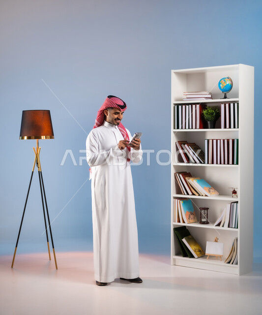 التواصل مع الأصدقاء والأقارب عبر التطبيقات والتقنيات الحديثة والمتطورة, القيام بالبث المباشر عبر السوشال ميديا, بورتريه لرجل عربي خليجي سعودي يرتدي الثوب التقليدي والشماغ يستخدم الهاتف المحمول, صورة الجسم بالطول الكامل, خلفية ملونة